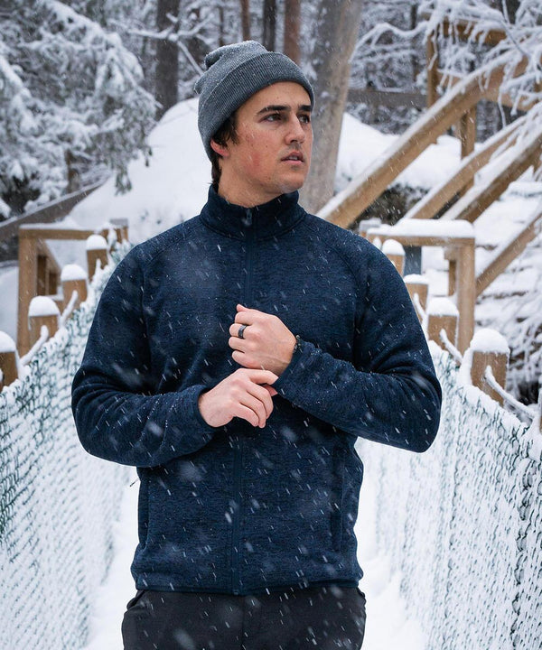 A man in a grey beanie and Stormtech Yosemite full-zip fleece jacket stands on a snowy bridge amid falling snow and trees, looking to the side with one hand touching his collar.