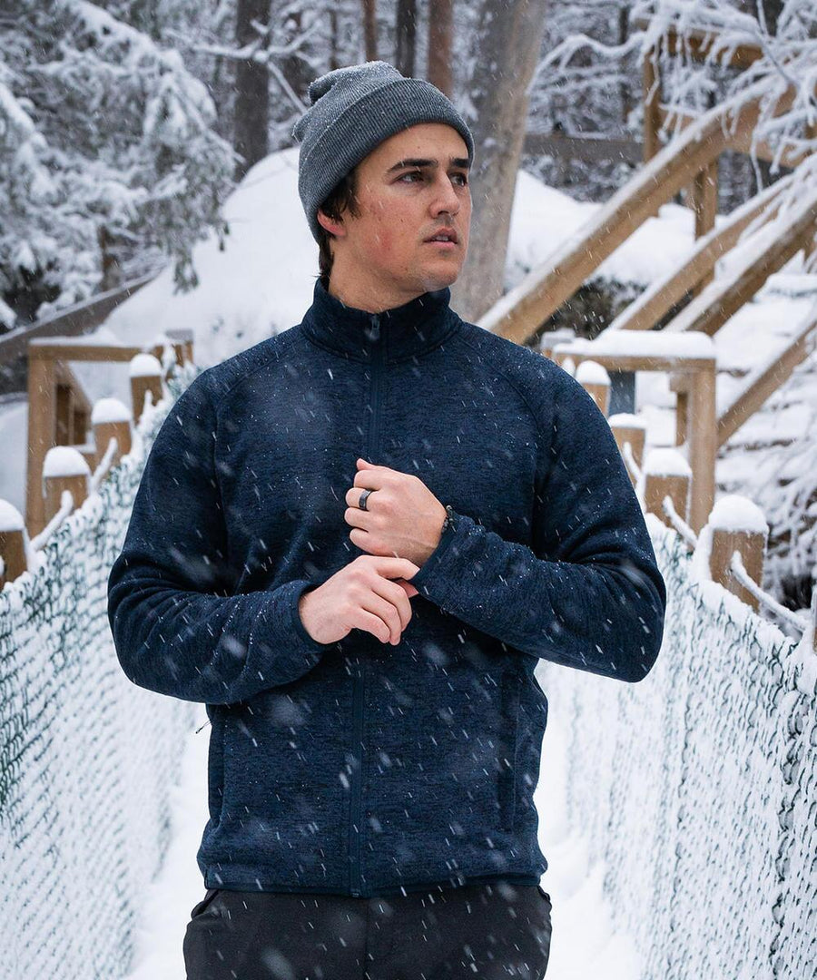 A man in a grey beanie and Stormtech Yosemite full-zip fleece jacket stands on a snowy bridge amid falling snow and trees, looking to the side with one hand touching his collar.