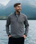 A man stands by a lake with mountains behind him, wearing the Stormtech Yosemite 1/4-zip pullover and dark trousers, looking to the side with his hands in his pockets.