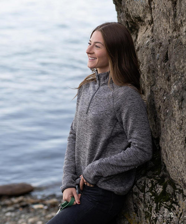 A woman wearing the Stormtech Women's Yosemite ¼-zip pullover in grey smiles as she leans against a rocky wall near water, holding something green, with rocks and a blurred shoreline in the background.