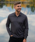 A man with short light brown hair and a beard stands by a calm lake, wearing the Stormtech Monterey 1/4-zip pullover in dark grey and black trousers, with one hand in his pocket as trees reflect on the water behind him.