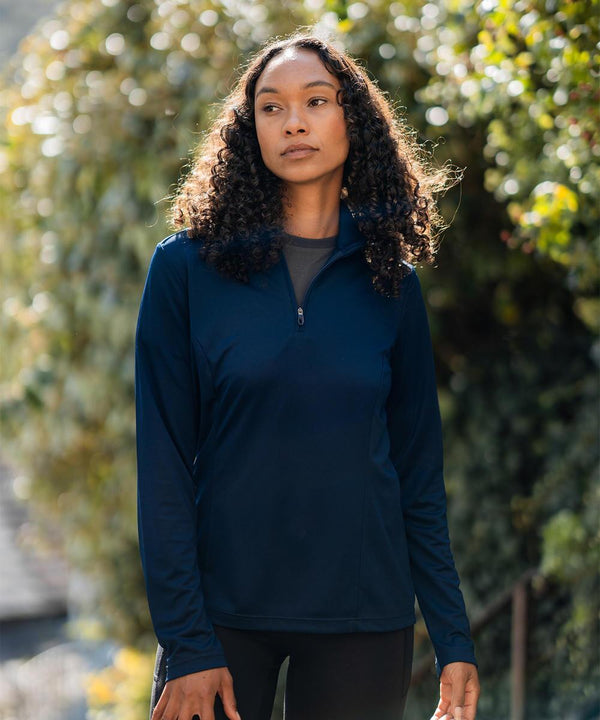 A person with long curly hair is standing outdoors, wearing the Stormtech Women's Monterey 1/4-zip pullover in navy blue and black trousers, with green foliage and soft sunlight in the background.