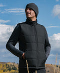 A man wears the Stormtech Sierra thermal gilet over a long-sleeve shirt and grey beanie, standing outdoors with his hands in the gilet pockets, looking to the side with clouds and trees in the background.