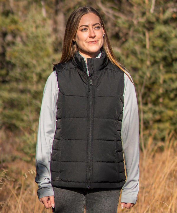 A woman stands outdoors in front of trees and dry grass, smiling as she wears the Stormtech Women’s Sierra thermal gilet over a light grey long-sleeve shirt and dark trousers.