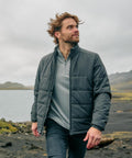 A man with brown hair and a beard wears the Stormtech Sierra thermal jacket and jeans whilst walking outdoors by water, with mountains and cloudy skies in the background.