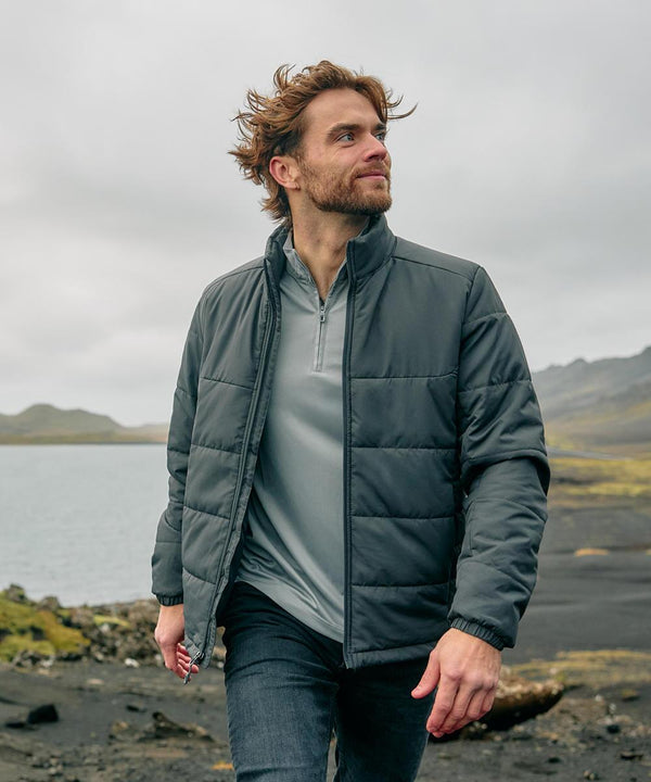 A man with brown hair and a beard wears the Stormtech Sierra thermal jacket and jeans whilst walking outdoors by water, with mountains and cloudy skies in the background.