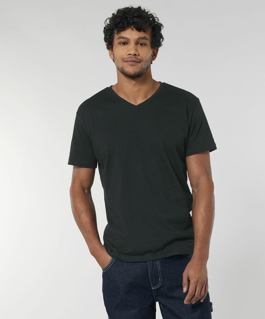 A man with curly hair is wearing a black V-neck T-shirt (SX021) by Stanley/Stella and blue jeans against a plain background.