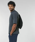 A man with curly hair, wearing a gray T-shirt and blue pants, carries a Stanley/Stella SX060 black backpack over one shoulder and looks towards the camera against a plain background.