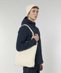A person wearing a navy jacket and a beige beanie hat stands holding a Stanley/Stella SX061 tote bag over their shoulder.
