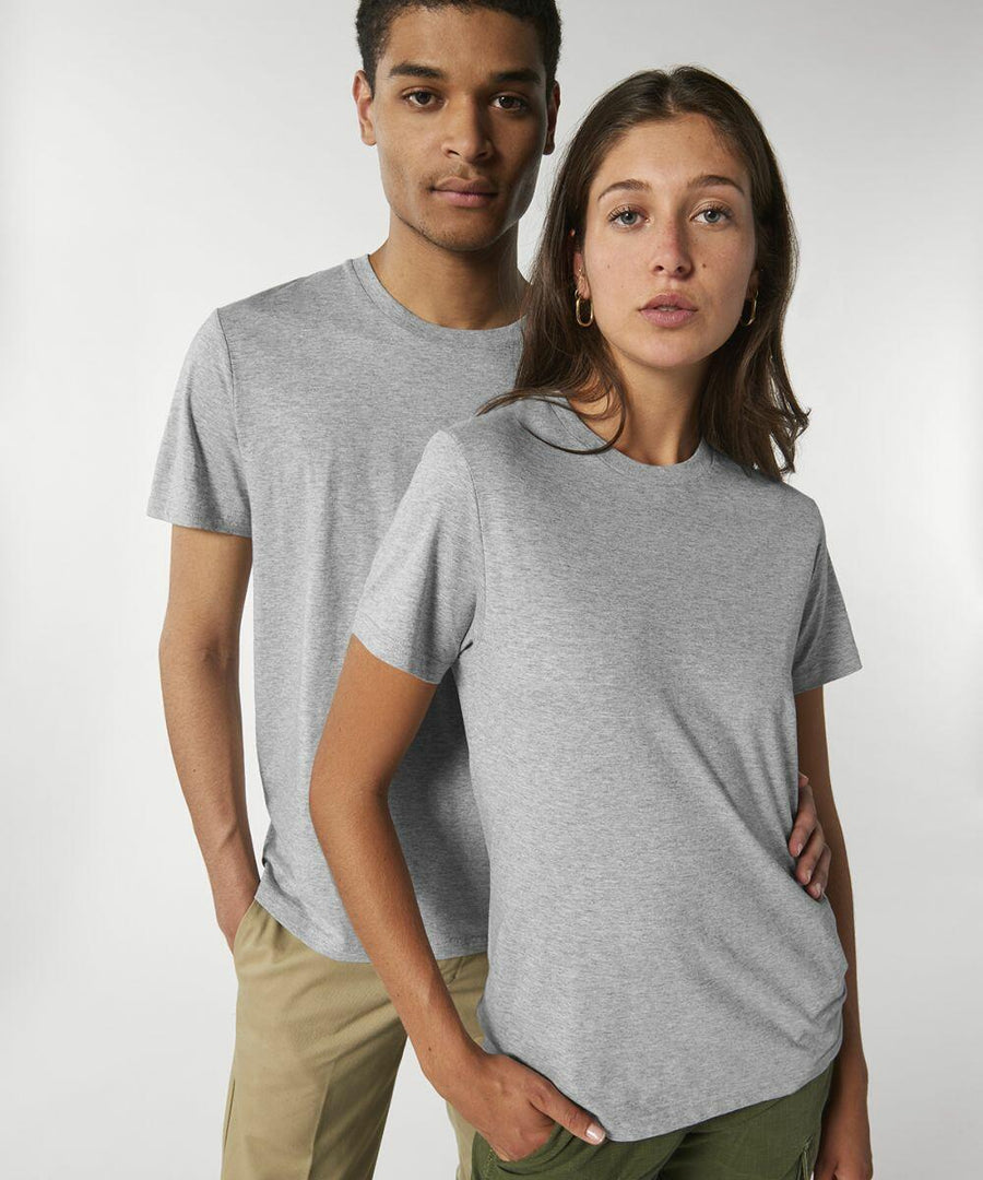 Two individuals stand side by side, both wearing grey SX087 t-shirts from Stanley/Stella and light-colored pants. The person in front has their hand in the pocket while the other rests their arm on the shoulder of the person in front.