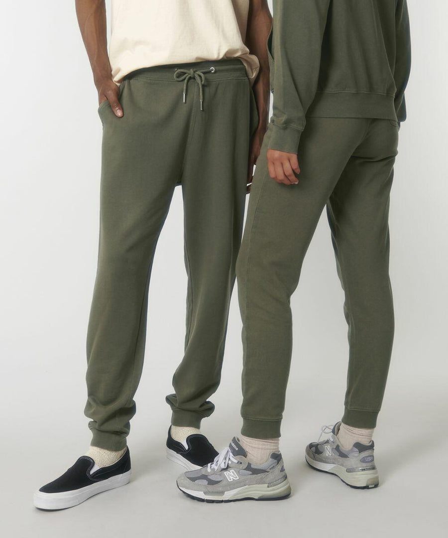 Two individuals wearing olive green sweatpants and casual footwear. One is wearing slip-on shoes and the other is wearing sneakers. Their upper clothing, Stanley/Stella SX100, is partially visible.