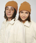Two individuals wearing Stanley/Stella SX146 brown beanies and light-colored jackets stand close to each other, both smiling gently. The person on the left has dreadlocks and freckles, while the person on the right has straight hair.