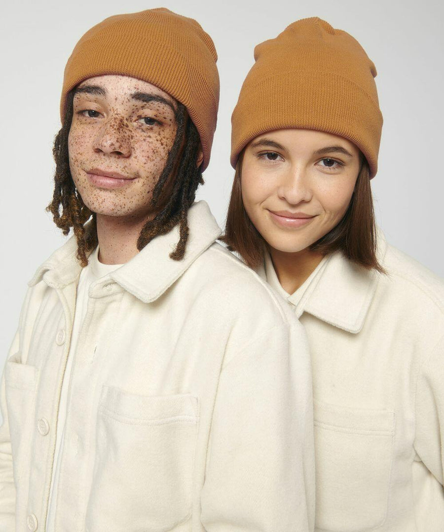 Two individuals wearing Stanley/Stella SX146 brown beanies and light-colored jackets stand close to each other, both smiling gently. The person on the left has dreadlocks and freckles, while the person on the right has straight hair.