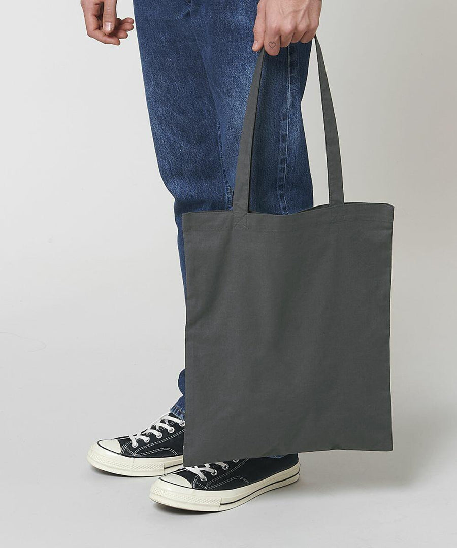 Person wearing blue jeans and black sneakers, holding a dark gray Stanley/Stella SX148.