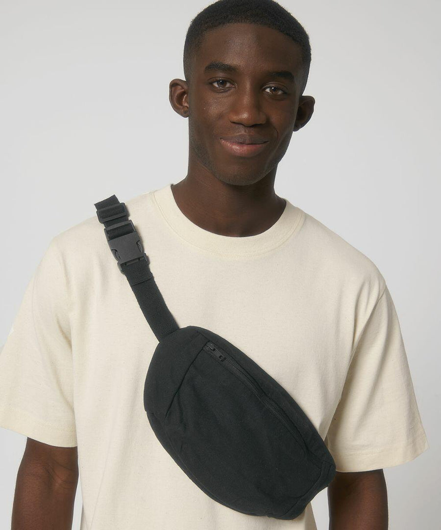 A man wearing a beige Stanley/Stella SX161 t-shirt and carrying a black sling bag looks at the camera against a plain background.