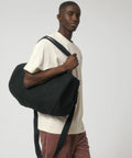 A person with a short haircut is wearing a light beige t-shirt and brown pants, carrying a large black Stanley/Stella SX162 over their shoulder against a plain white background.