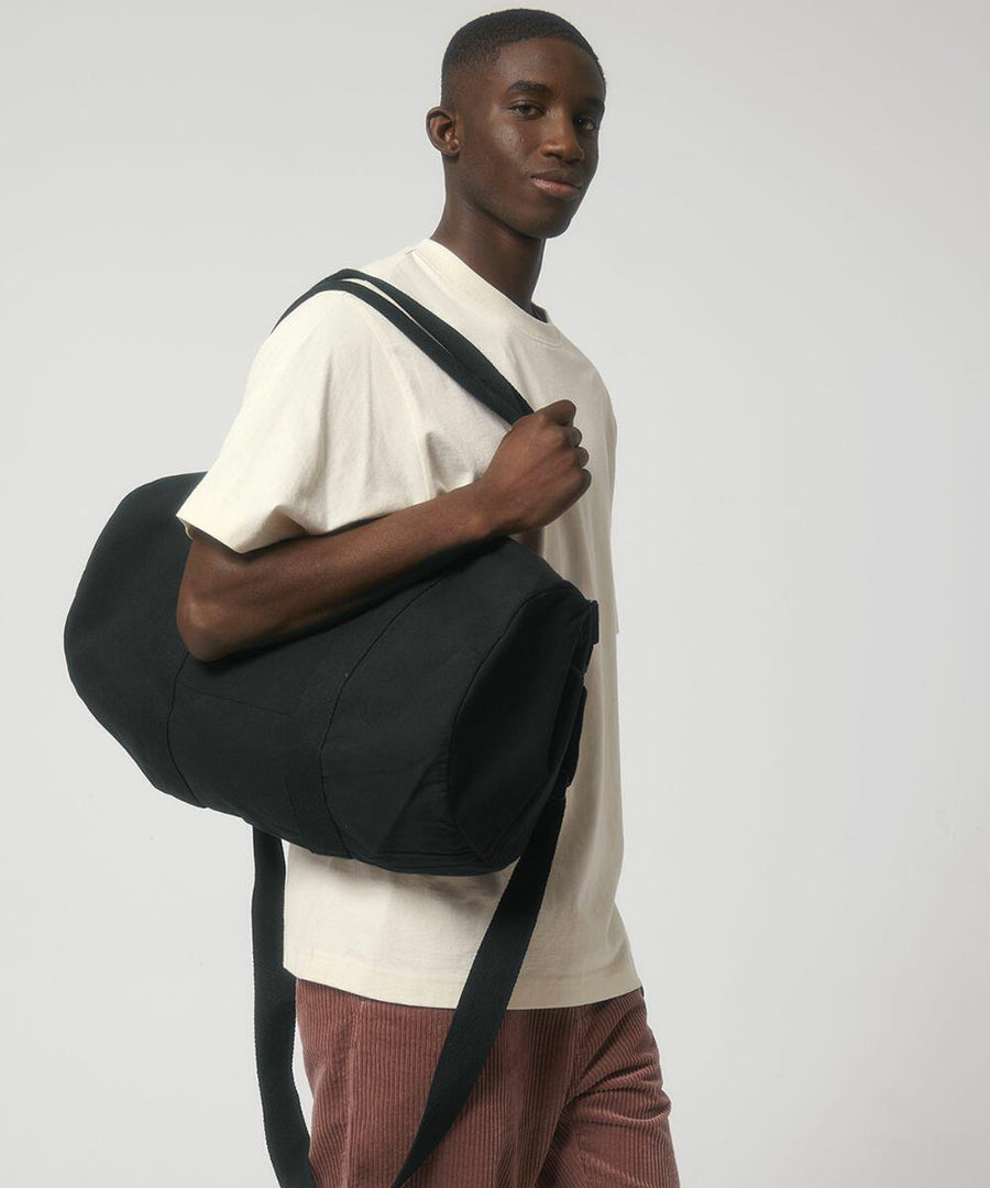 A person with a short haircut is wearing a light beige t-shirt and brown pants, carrying a large black Stanley/Stella SX162 over their shoulder against a plain white background.
