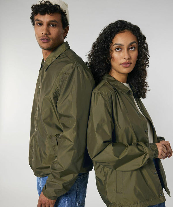 Two individuals, one with short curly hair and the other with long curly hair, both wearing green SX176 jackets by Stanley/Stella and standing back-to-back, facing the camera against a plain background.