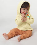A toddler in a yellow hoodie and brown pants sits on the floor, chewing on a colorful SX186 by Stanley/Stella.