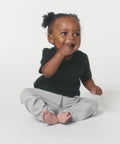 A small child with curly hair in a black t-shirt and gray pants sits on the floor, touching their mouth and smiling while playing with the SX194 from Stanley/Stella.