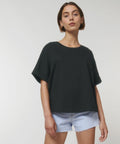 A person with short brown hair is wearing a loose black Stanley/Stella SX198 T-shirt and light blue denim shorts. They are standing and looking directly at the camera with a neutral expression.