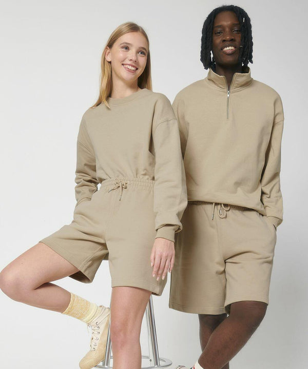 Two people standing side by side, both wearing matching SX200 beige outfits from Stanley/Stella with long-sleeved tops and shorts, and smiling at the camera. One person is on a stool while the other stands.