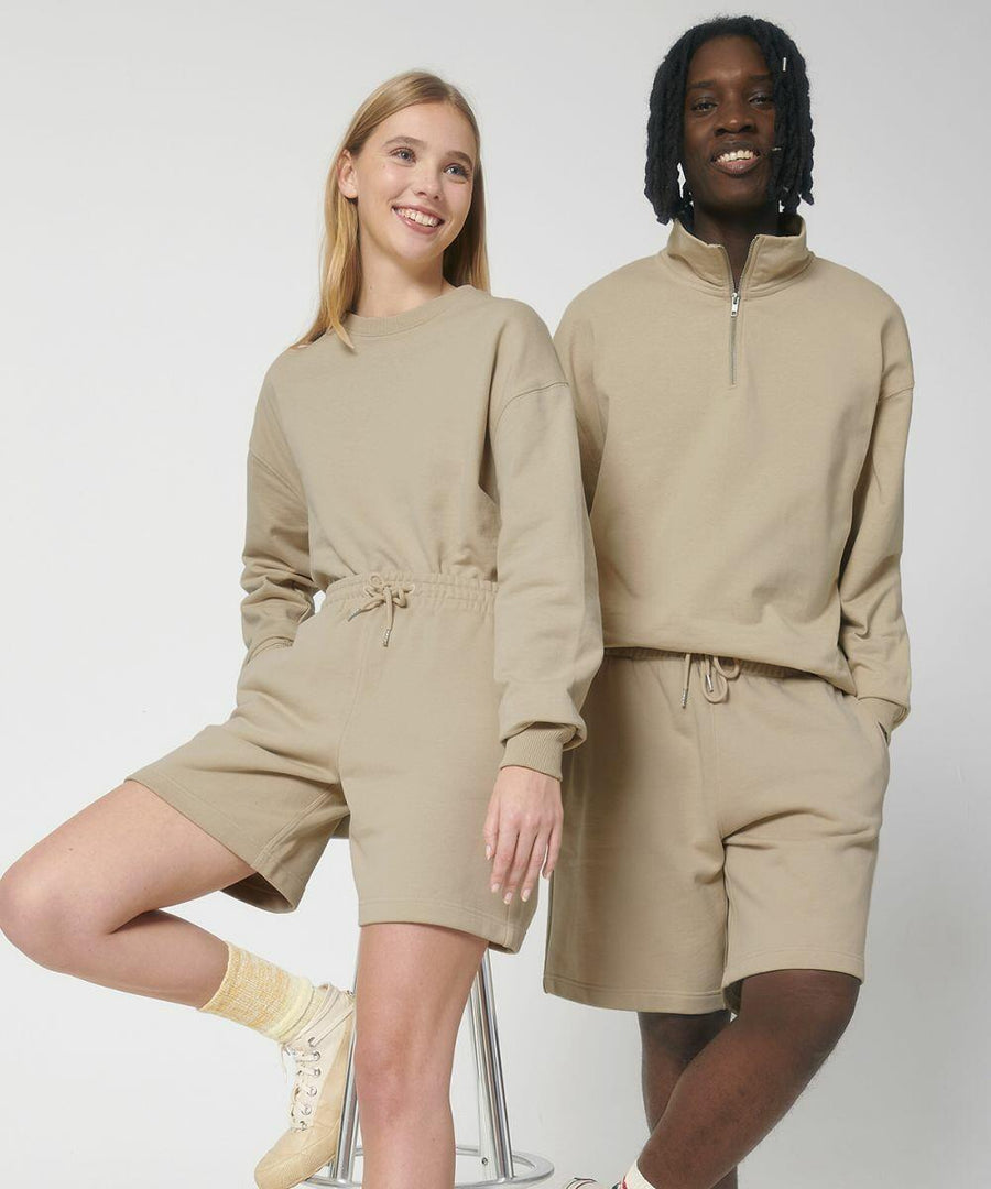 Two people standing side by side, both wearing matching SX200 beige outfits from Stanley/Stella with long-sleeved tops and shorts, and smiling at the camera. One person is on a stool while the other stands.