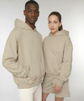 A man and a woman wearing Stanley/Stella SX217 beige hoodies pose together against a plain white background. The woman also wears beige shorts.