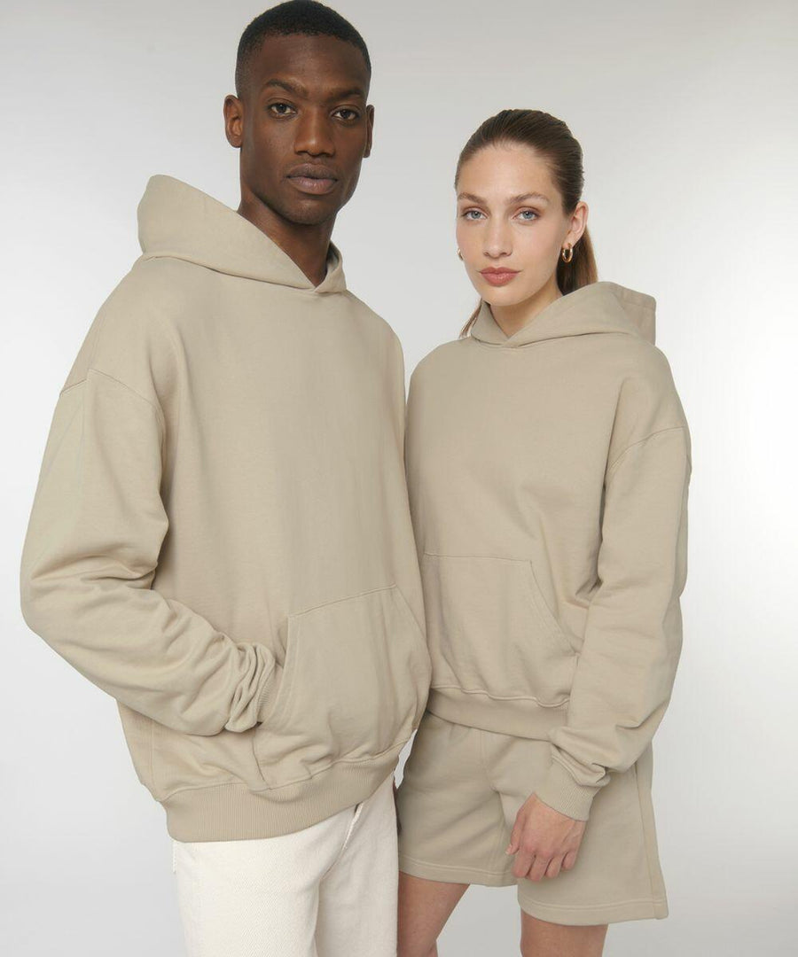 A man and a woman wearing Stanley/Stella SX217 beige hoodies pose together against a plain white background. The woman also wears beige shorts.