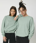 Two people wearing matching light green sweatshirts and black pants, with the person on the right resting an arm on the other’s shoulder. They both have neutral expressions and are standing against a plain background, showcasing the Stanley/Stella SX218.