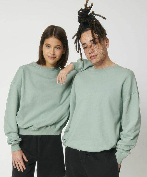 Two people wearing matching light green sweatshirts and black pants, with the person on the right resting an arm on the other’s shoulder. They both have neutral expressions and are standing against a plain background, showcasing the Stanley/Stella SX218.