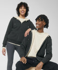Two individuals wearing dark SX222 zip-up hoodies from Stanley/Stella and light-colored t-shirts, smiling, against a white background. One stands while the other sits on a stool.
