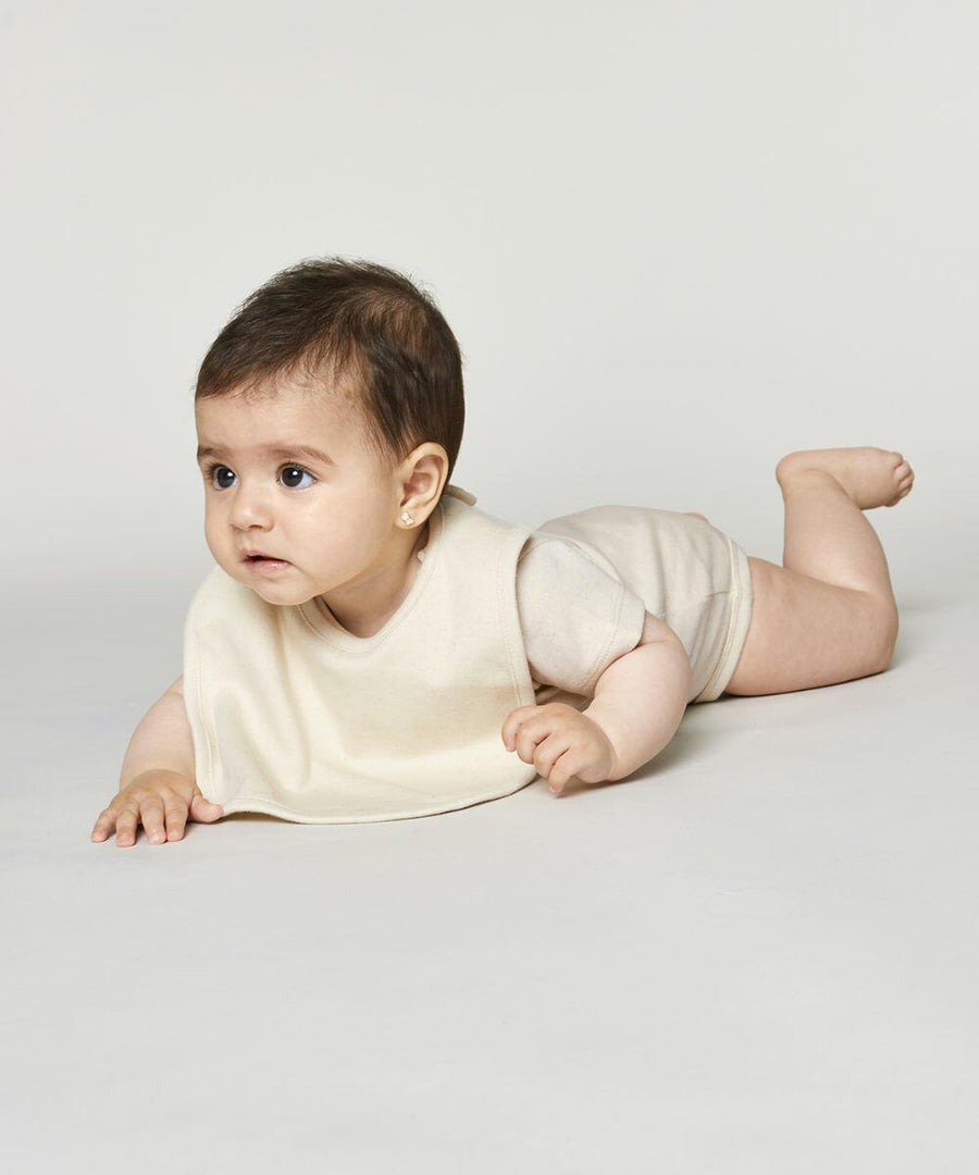 A baby in a beige SX227 Stanley/Stella onesie and bib lies on their stomach on a plain white background.