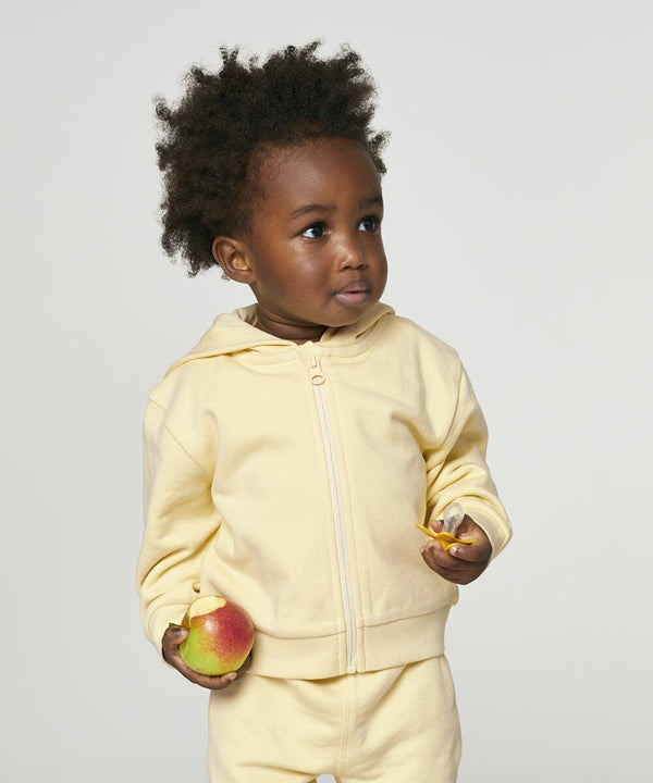 A toddler wearing a yellow jacket and pants holds a red and yellow apple and a bitten piece of fruit, standing against a plain white background, showcasing the SX229 by Stanley/Stella.