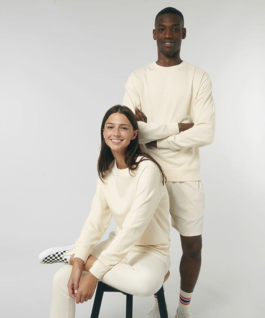 Two people are posing in off-white casual outfits. The person seated is wearing a Stanley/Stella SX230 long-sleeve shirt and pants, while the person standing is wearing a Stanley/Stella SX230 long-sleeve shirt and shorts.