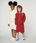 Two children stand together. One wears a beige SX232 by Stanley/Stella hoodie and shorts, while the other wears a red SX232 by Stanley/Stella hoodie and matching shorts. Both are smiling and have their arms around each other.