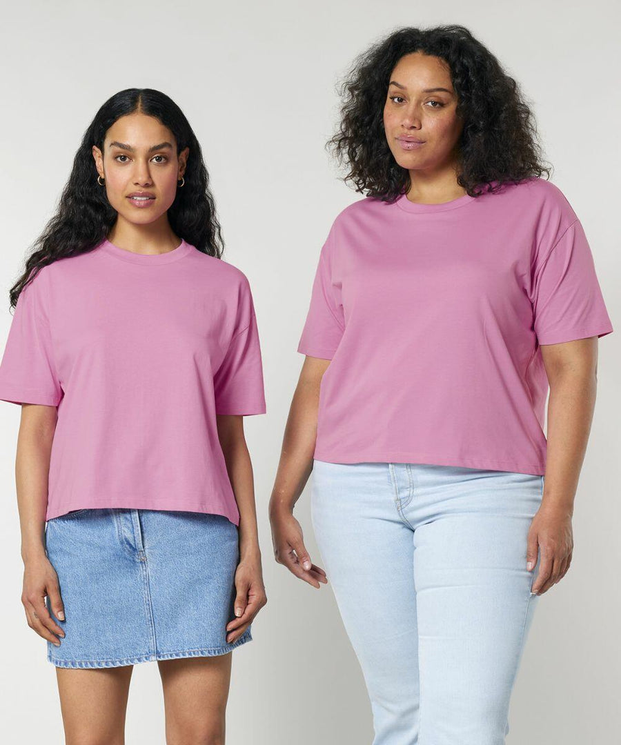 Two individuals wearing similar pink SX235 t-shirts by Stanley/Stella. The person on the left wears a blue denim skirt, while the person on the right wears light blue jeans. Both standing against a plain background.