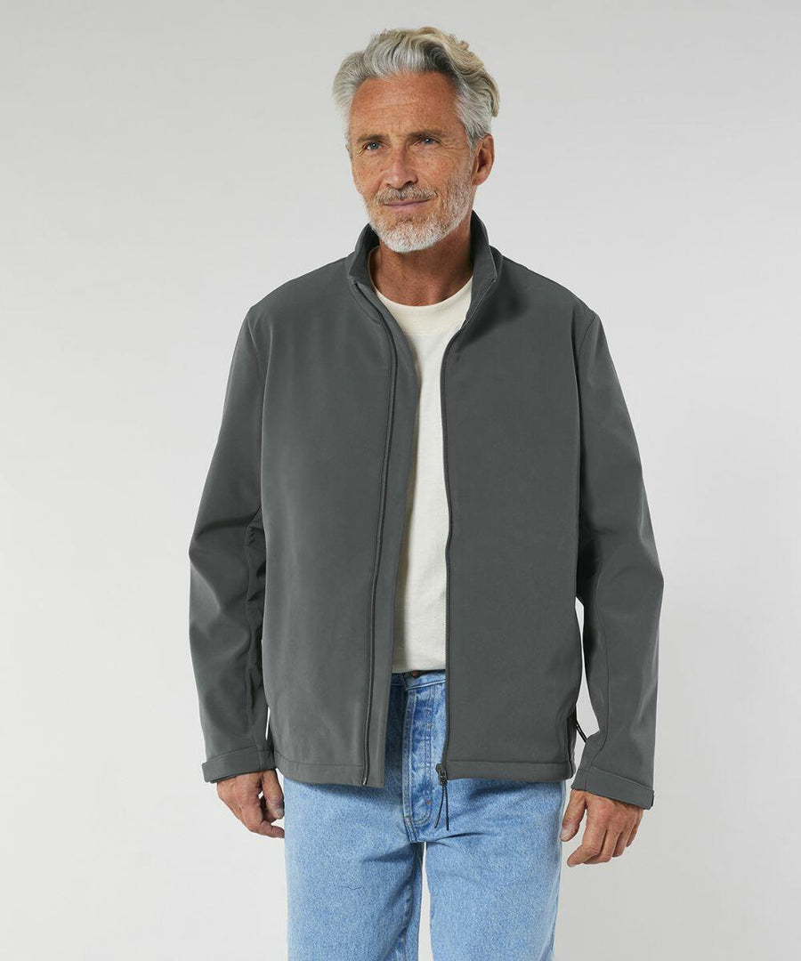 A man with gray hair and beard wearing a gray Stanley/Stella SX240 jacket over a white shirt and blue jeans stands against a plain background.