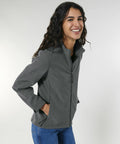 Person with long hair wearing a gray Stanley/Stella SX241 zip-up jacket and blue jeans, smiling and posing against a plain background.