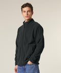 A man stands facing forward wearing the Stanley/Stella Stanley Guider (STJM238) black zip-up fleece jacket with a high collar and blue jeans. His left hand is in his pocket, looking slightly aside against a plain, light background.