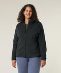 A woman with curly hair wears the Stanley/Stella Stella Guider (STJW239) black zip-up jacket and blue jeans, standing and smiling against a plain light background with her hands relaxed at her sides.