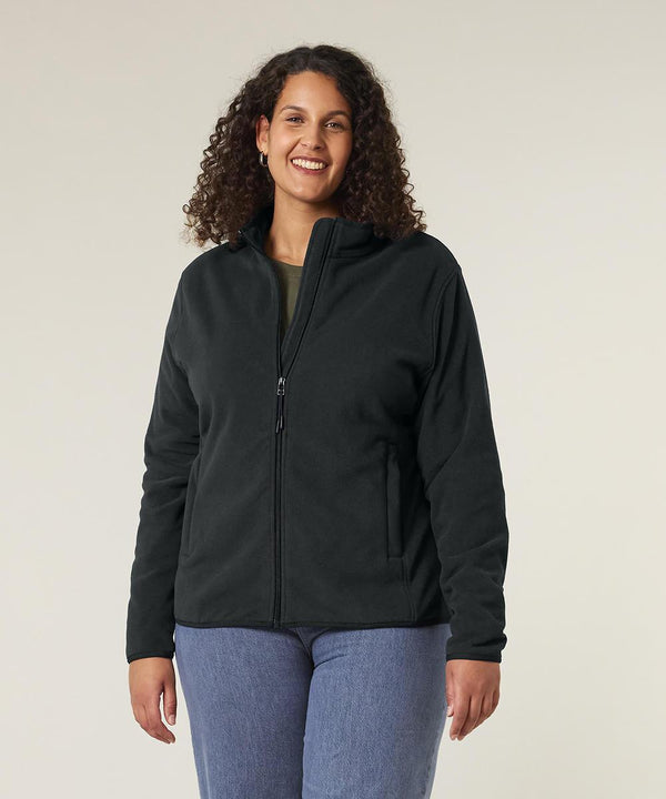 A woman with curly hair wears the Stanley/Stella Stella Guider (STJW239) black zip-up jacket and blue jeans, standing and smiling against a plain light background with her hands relaxed at her sides.