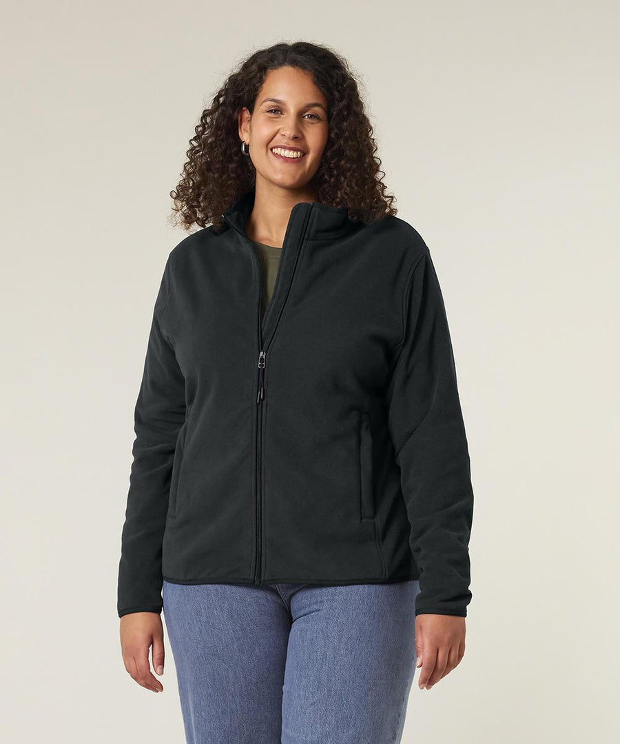 A woman with curly hair wears the Stanley/Stella Stella Guider (STJW239) black zip-up jacket and blue jeans, standing and smiling against a plain light background with her hands relaxed at her sides.