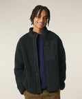 A person faces the camera, smiling, dressed in a Stanley/Stella Brooker (STJU248) black fleece jacket with a high collar and zipped chest pocket over a dark shirt and light brown trousers. The background is plain and light-coloured.