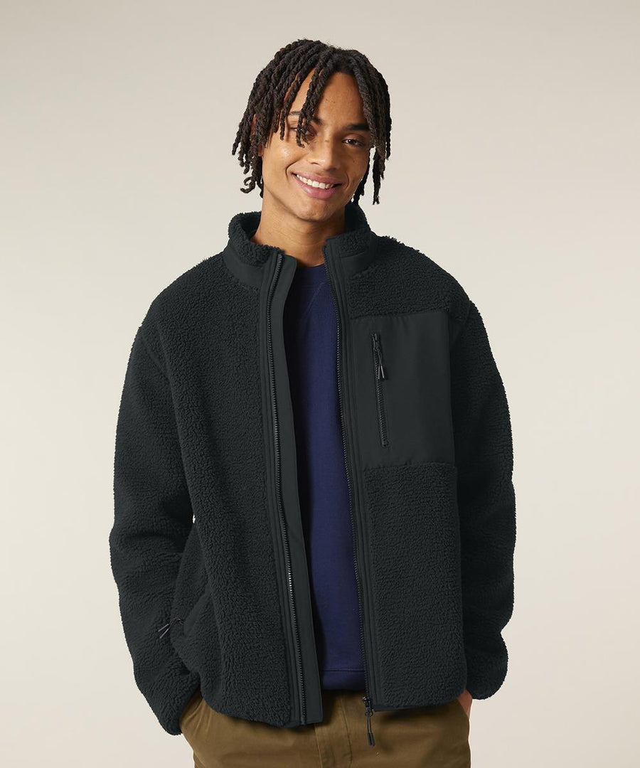 A person faces the camera, smiling, dressed in a Stanley/Stella Brooker (STJU248) black fleece jacket with a high collar and zipped chest pocket over a dark shirt and light brown trousers. The background is plain and light-coloured.