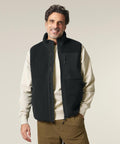 A man with wavy brown hair and a beard is smiling at the camera, wearing the Stanley/Stella Liner (STJU249) black fleece zip-up gilet over a cream sweatshirt and brown trousers, standing against a light grey background.