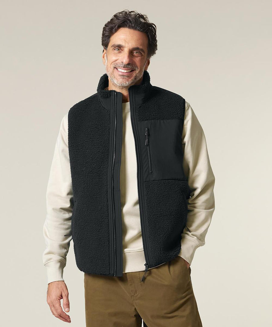 A man with wavy brown hair and a beard is smiling at the camera, wearing the Stanley/Stella Liner (STJU249) black fleece zip-up gilet over a cream sweatshirt and brown trousers, standing against a light grey background.