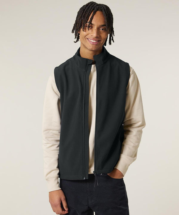 A person wears the Stanley Trail blazer (STJM235) by Stanley/Stella—a light beige long-sleeve shirt layered with a black zip-up gilet and dark trousers—standing against a plain light background, smiling with one hand in their pocket.