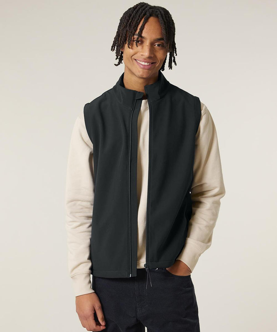 A person wears the Stanley Trail blazer (STJM235) by Stanley/Stella—a light beige long-sleeve shirt layered with a black zip-up gilet and dark trousers—standing against a plain light background, smiling with one hand in their pocket.