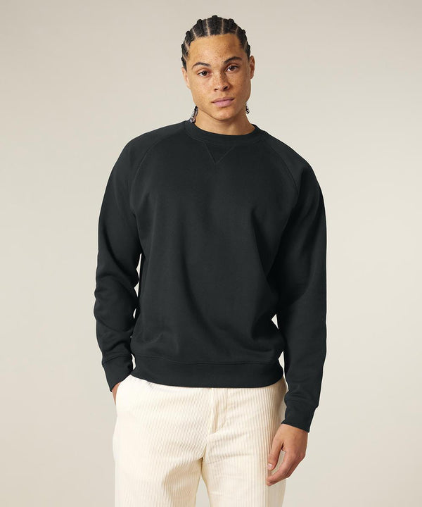 A person wears a black Stanley/Stella Knoxer (STSU201) crewneck sweatshirt and light trousers, standing facing forward against a plain, light background.